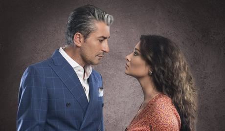 Erkan Petekkaya wearing a blue suit and a white shirt while facing Nurgül Yeşilçay with curly long hair, wearing earrings and an orange dress.