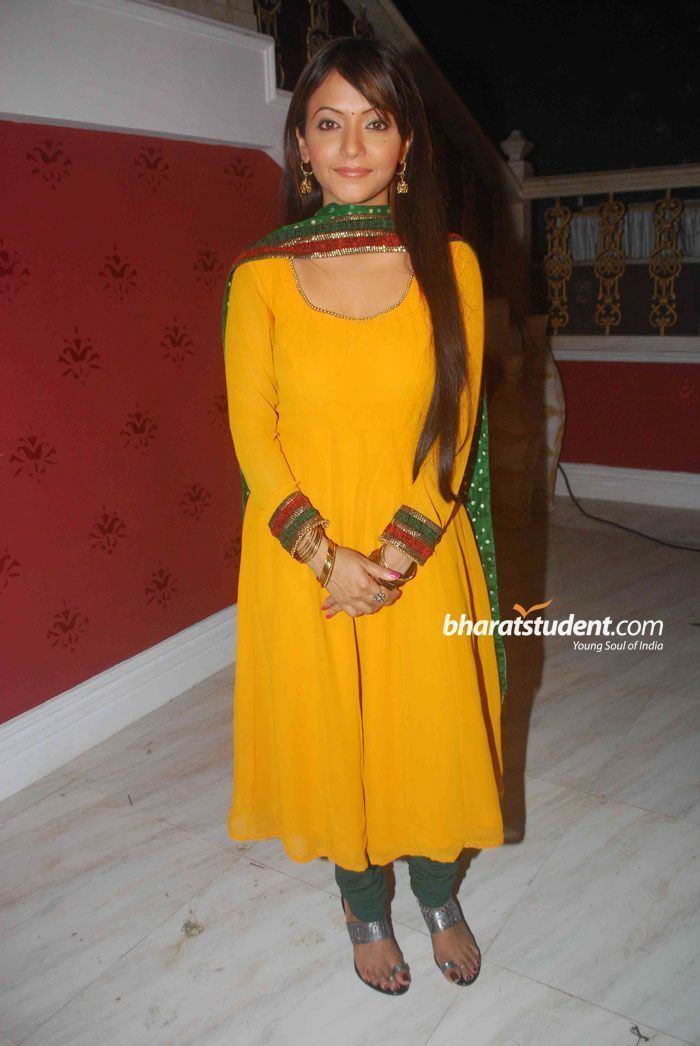 Pariva Pranati smiling while wearing a yellow dress, green and red scarf, green pants, gray sandals, bracelet, and earrings