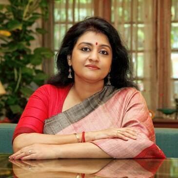 Parvathy Jayaram is smiling, has black hair and a bindi, and sitting down on a blue chair, she is wearing silver earrings, a bracelet on her right hand, and a red blouse with a black-red saree.