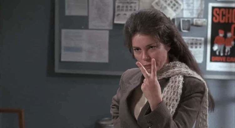 Pat Quinn (actress) showing her two fingers while wearing a brown coat and a scarf inside a room in a movie scene from Alice's Restaurant (1969)