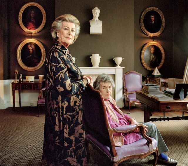 Countess Patricia Knatchbull sitting on a chair while Lady Pamela Hicks standing on her back, both with serious faces while looking at the camera. Countess Patricia wearing an off-white vest over a pink long sleeve blouse, and gray pants while Lady PamelaLady Pamela wearing a floral black dress.