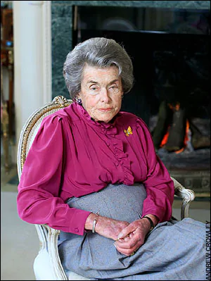 Old Patricia Knatchbull with a serious face while sitting on a white chair, wearing a magenta long sleeve blouse and a gray skirt.