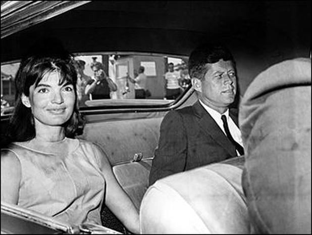 Jacqueline Kennedy smiling while inside the car, wearing a sleeveless dress, and Patrick Bouvier Kennedy sitting beside her