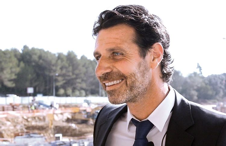 Patrick Mouratoglou with a smiling face, wearing a black suit, black tie, and white long sleeves.