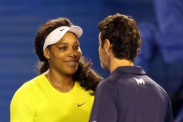 Patrick Mouratoglou wearing a blue polo shirt together with Serena Williams with a smiling face, wearing a white cap, a necklace, and a yellow shirt.
