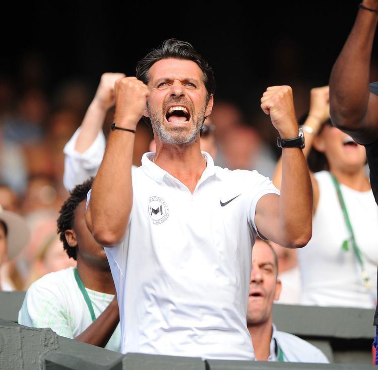 Patrick Mouratoglou wearing a watch, wristband, a white polo shirt while screaming for victory.
