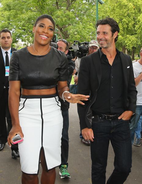 Patrick Mouratoglou with a smiling face, wearing a black suit, black shirt, and black pants with Serena Williams wearing a black top, white and black skirt, and a necklace while holding a phone and surrounded by media.