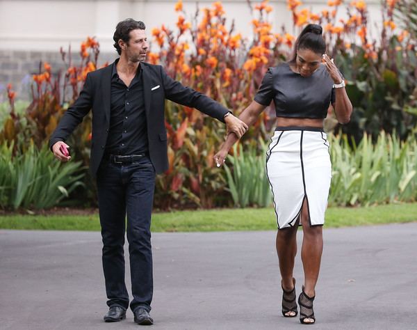 Patrick Mouratoglou wearing a black suit, black shirt, black pants, black belt, and black shoes while holding Serena Williams wearing a black top, white and black skirt, and black sandals.