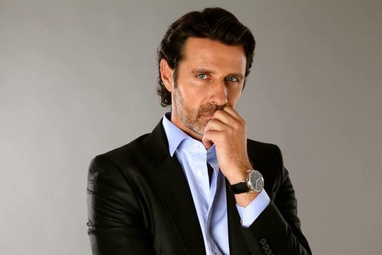 Patrick Mouratoglou with a serious face, wearing a black suit, light blue long sleeves, and a watch.