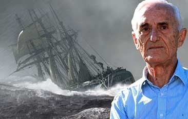 At the back is a black and white storm sky in a sea with high strong waves, an old boat is sinking from its bow has black mast , white jib, and black body, at the right, Patrick O'Brian is serious, standing, has white hair black eyebrow, wearing a blue polo shirt.