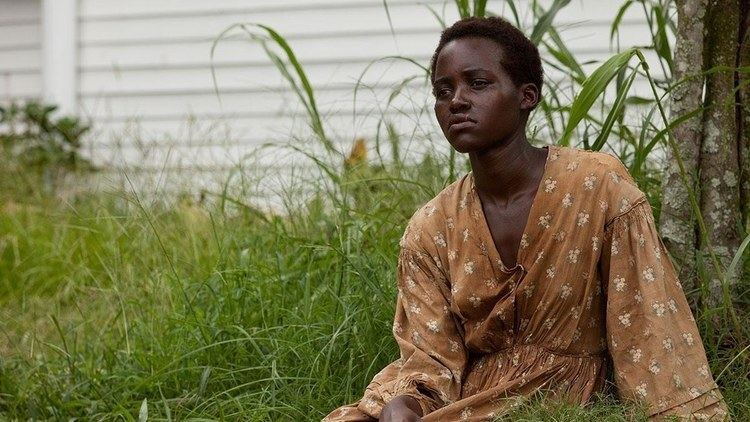 Lupita Nyong'o as Patsey, crying and looking afar while wearing a brown and white long sleeve dress in the 2013 film, 12 Years a Slave