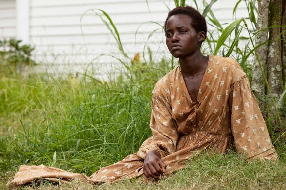 Lupita Nyong'o as Patsey, crying and looking afar while wearing a brown and white long sleeve dress in the 2013 film, 12 Years a Slave