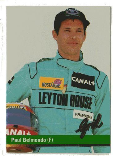 Paul Belmondo smiling while holding a helmet and wearing a black and blue racing suit and black cap