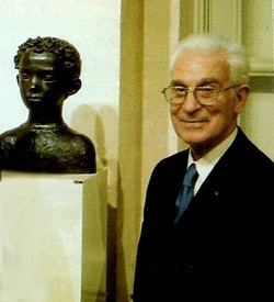 Paul Belmondo with a tight-lipped smile while standing beside the sculpture and wearing a black coat, white long sleeves, and necktie