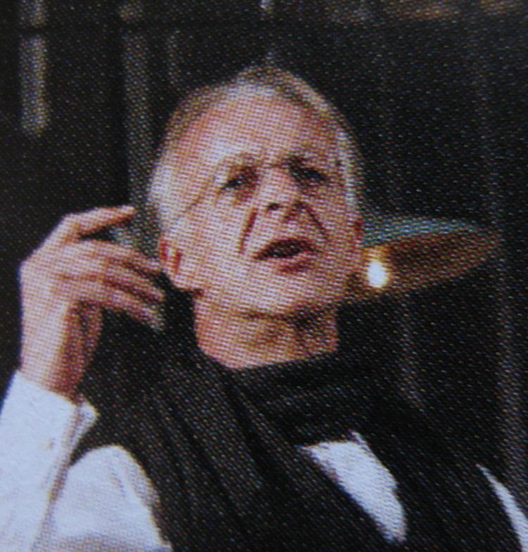 Paul Bown talking to someone while wearing white long sleeves, black scarf, and eyeglasses