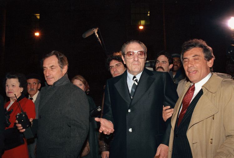 Paul Castellano in the middle of media and paparazzi wearing a black long coat as well as red tinted glasses.