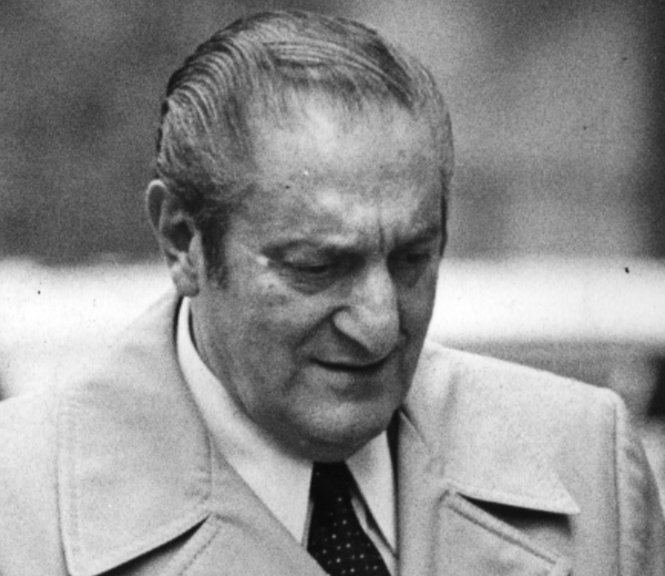 Paul Castellano with streaks of gray hair wearing a long coat as well as a black spotted tie.