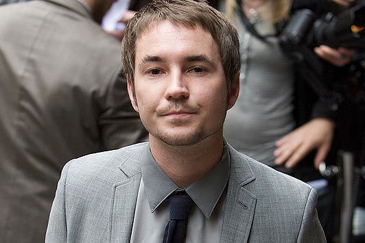 Martin Compston with a tight-lipped smile while wearing a gray coat, gray long sleeves, and black necktie