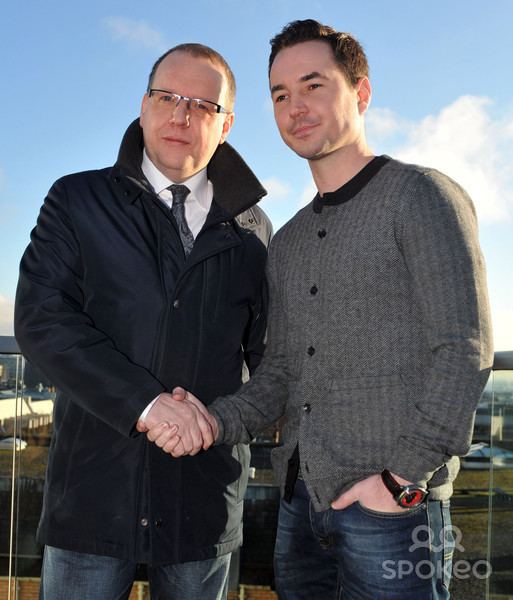 Paul Ferris shaking hands with Martin Compston while he is wearing a black jacket, white long sleeves, black necktie, and denim pants