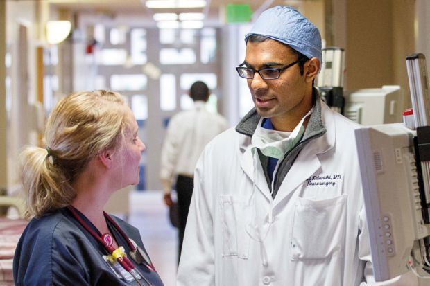 Paul Kalanithi talking to his colleague at the Stanford hospital while wearing a surgical cap, eyeglasses, and lab gown