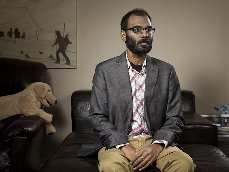 Paul Kalanithi sitting on a black couch while looking afar and wearing a gray coat, checkered long sleeves, brown pants, and eyeglasses