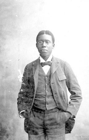 Paul Laurence Dunbar at age 19, 1892, looking at something while hands are in his pocket, with a serious face, and wearing eyeglasses, pants, and a long sleeve under a vest, bow tie, and coat