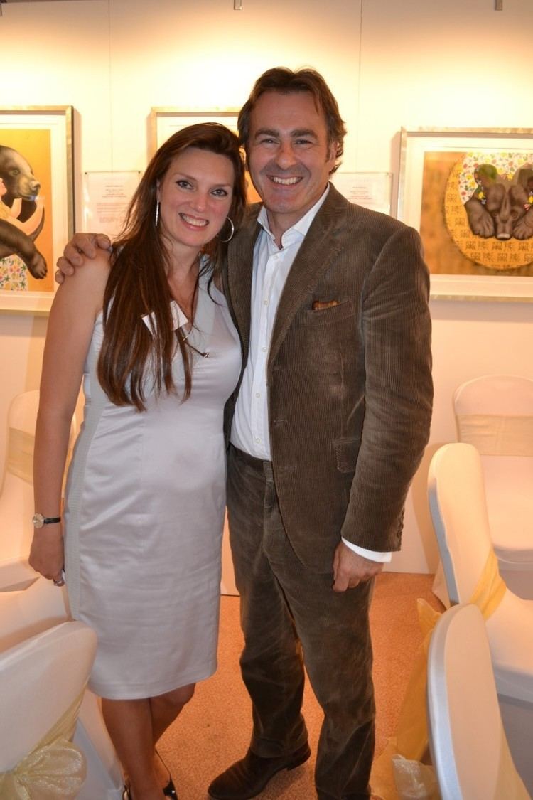 Paul Martin wearing a brown suit, a white shirt, and brown pants with his wife, Charlotte Godfrey with long hair, wearing earrings, a watch, and a sexy white dress.