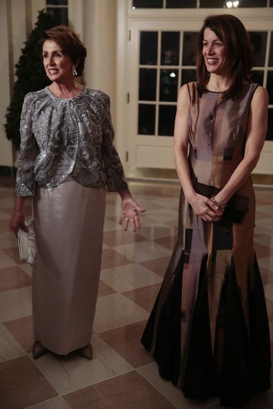 Nancy Pelosi wearing earrings, a fancy dress, and holding a white pouch with Jacqueline Pelosi wearing a brown sleeveless gown and holding a brown pouch.