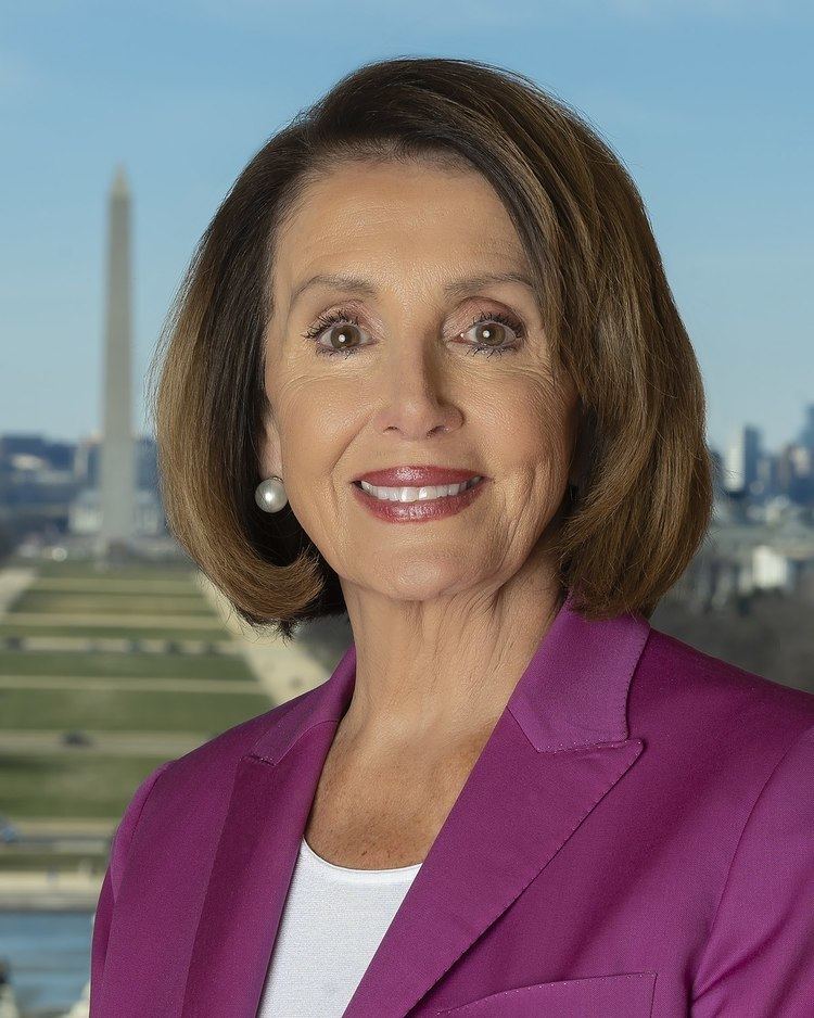 Nancy Pelosi with short blonde hair, wearing earrings, and a purple suit.