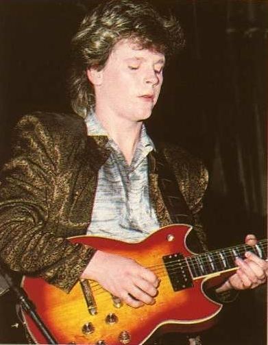 Paul Reynolds playing guitar while wearing a brown blazer and gray long sleeves