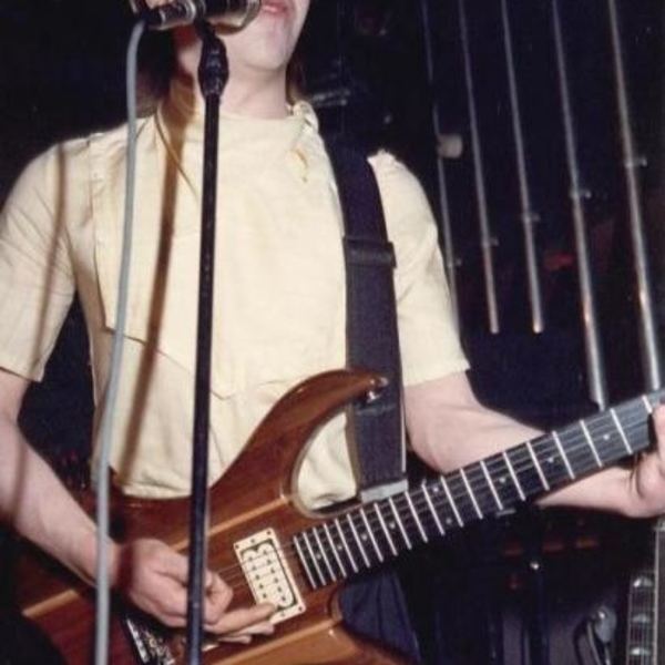 Paul Reynolds playing guitar while singing and wearing a yellow shirt and black pants