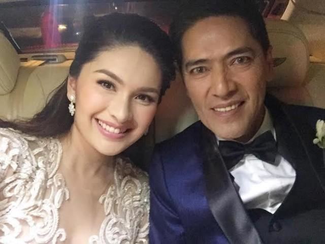 Pauleen Luna with smiling face, wearing earrings and a white wedding gown together with her husband Vic Sotto inside a car, wearing a blue suit and a black bow tie.