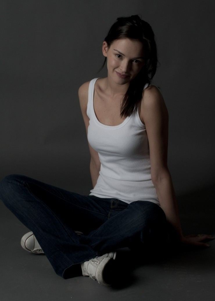 Paulina Andreeva with a tight-lipped smile while sitting on the floor and wearing a white tank top, denim pants, and white shoes
