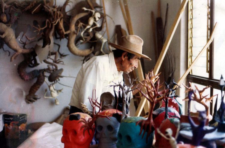 Pedro Linares standing and holding a plank of wood while surrounded by sculptures