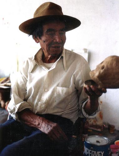 Pedro Linares sitting and wearing a white long sleeve and a hat while holding a skull sculpture
