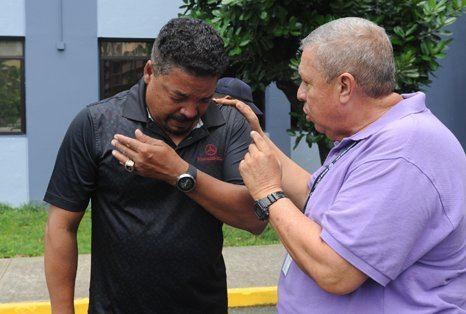 Pedro Rosa Nales is crying and the man beside him is talking to him while Pedro is wearing a black polo shirt, wristwatch, and ring