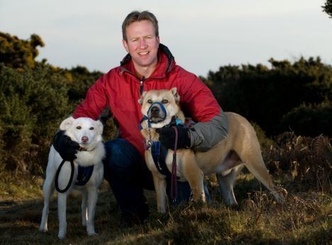 Pen Farthing smiling and wearing a red jacket while two dogs are beside him
