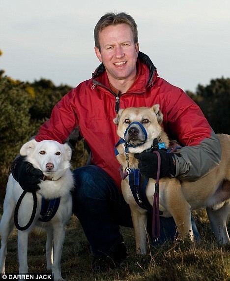 Pen Farthing smiling while wearing a red jacket with two dogs beside him