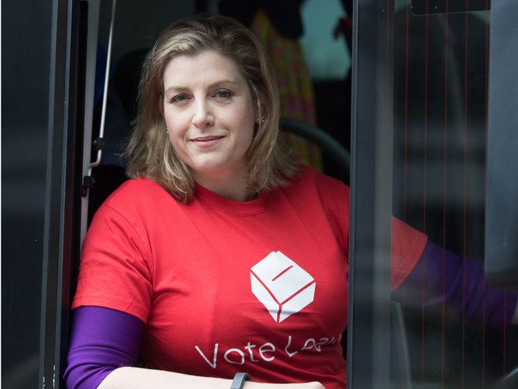 Penny Mordaunt is smiling, sitting in a bus, looking out at the vehicle window, has blond hair wearing a black bracelet, gold earrings, purple long sleeve under a red shirt with printed white logo, and a written word “VOTE”.