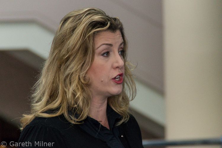 Penny Mordaunt is serious, looking to her left, mouth half open, eyebrows up, has wavy  blond hair, has mole in her right cheek,  wearing a black shirt.