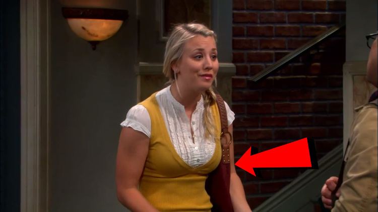 Kaley Cuoco as Penny talking to a man in front of her while her back is slouched and her long blonde hair tied sideways and behind her is a wall lantern and a brick wall with a staircase from a scene in a popular series, "The Big Bang Theory". She has a leather-strapped shoulder bag with a metal ring pairing it with a white buttoned blouse underneath her yellow sleeveless top.
