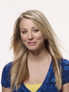 Kaley Cuoco smiling with her long blonde hair down and wearing a yellow shirt under a blue floral blouse.
