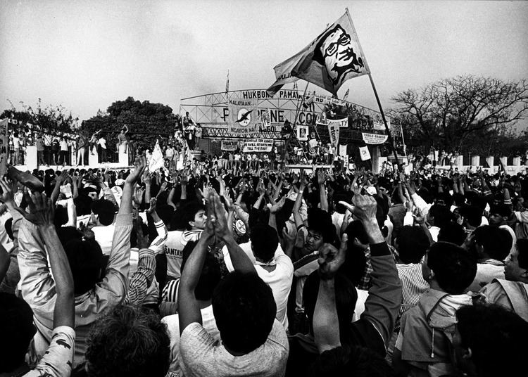 A crowd of people protesting doing an L hand gesture and carrying flags during the People Power Revolution