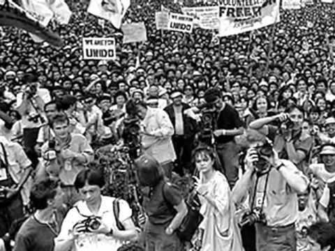A crowd of people protesting and carrying posters with written words against Marcos's reign and photographers taking pictures in the front  during the People Power Revolution