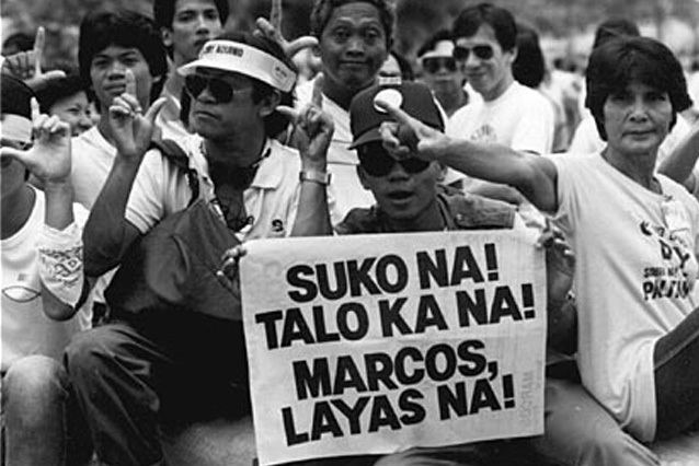 A crowd of people protesting doing an L hand gesture and carrying a poster with words written in Filipino meaning GIVE UP! YOU'VE LOST! MARCOS, GONE! against Marcos during the People Power Revolution