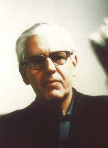 Peter Birkhäuser with a serious face, white hair, wearing eyeglasses, and a black coat over gray long sleeves.