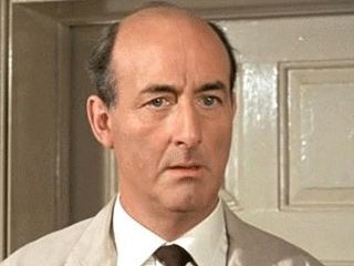 Peter Hughes in a movie scene wearing a cream colored coat over a white suit and dark necktie and a bald head.