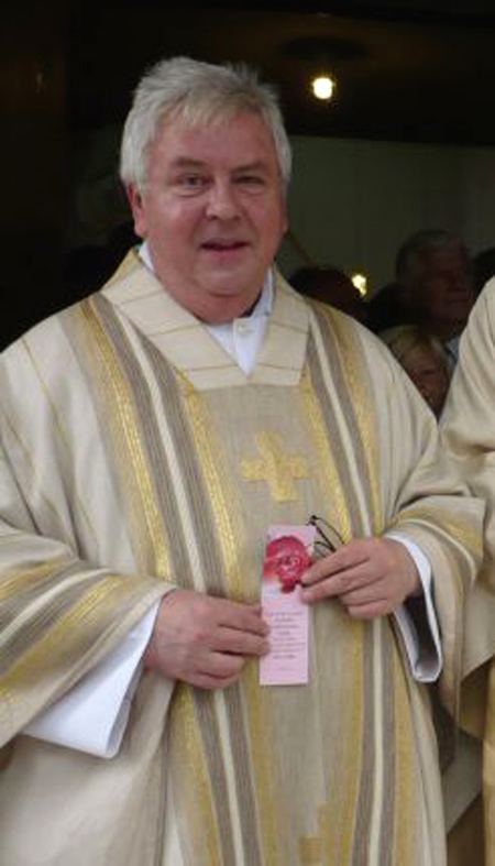 Peter Hullermann smiling while holding a pink pamphlet and eyeglasses while wearing a white long sleeve under a gold and beige chasuble with cross embroid on the front