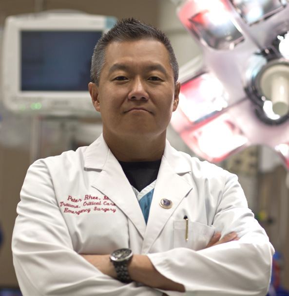 Peter M. Rhee posing with his arms crossed and wearing a lab gown inside an operating room.