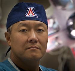 Peter M. Rhee smiling closed mouth inside an operating room and wearing a blue hat and a scrub.